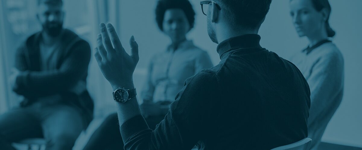Rear view of man with raised hand on group therapy in medical center. Rear view of man raising his hand to ask a question while attending group therapy.